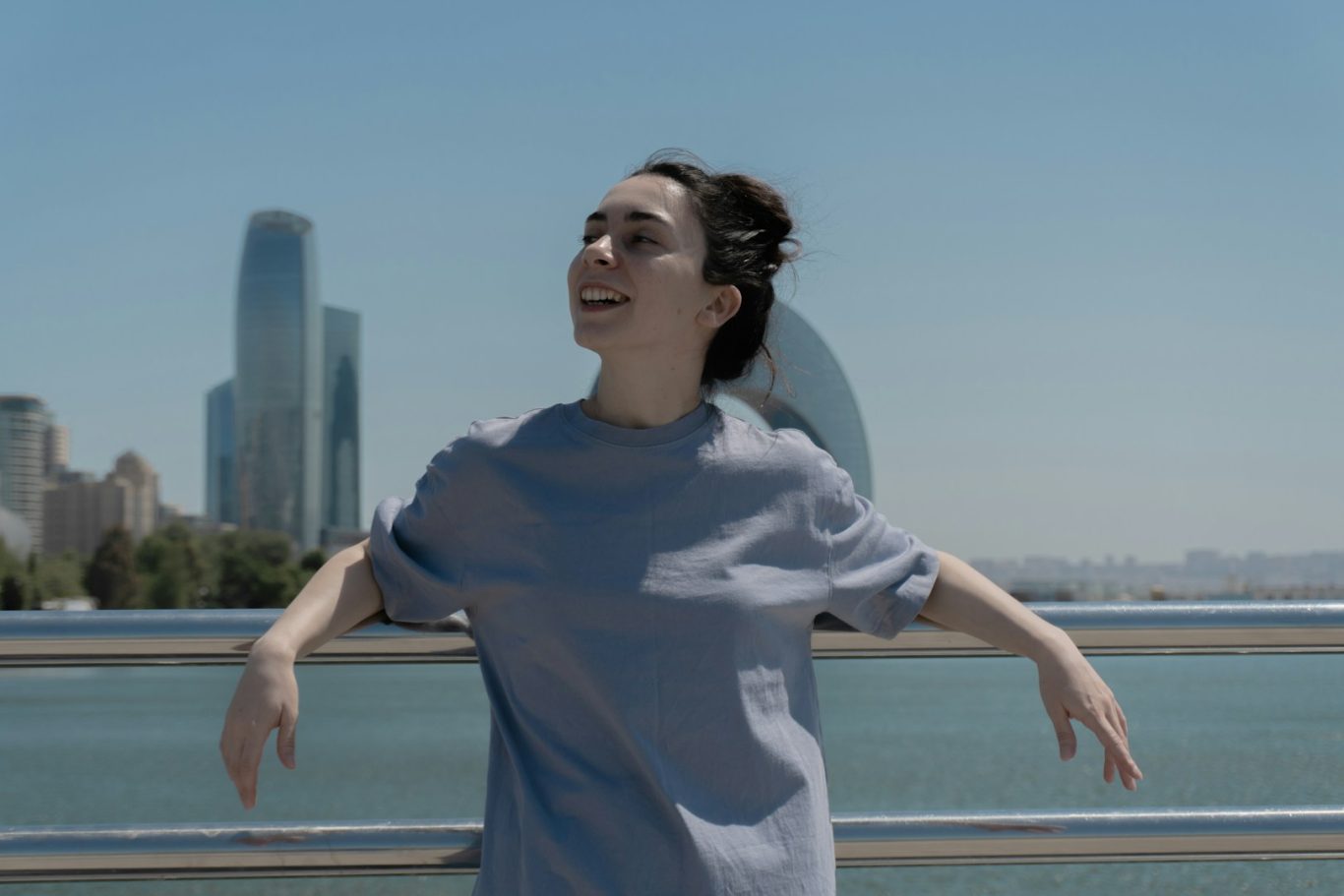 Giovane donna sorridente in maglietta grigia, con skyline urbano sullo sfondo.