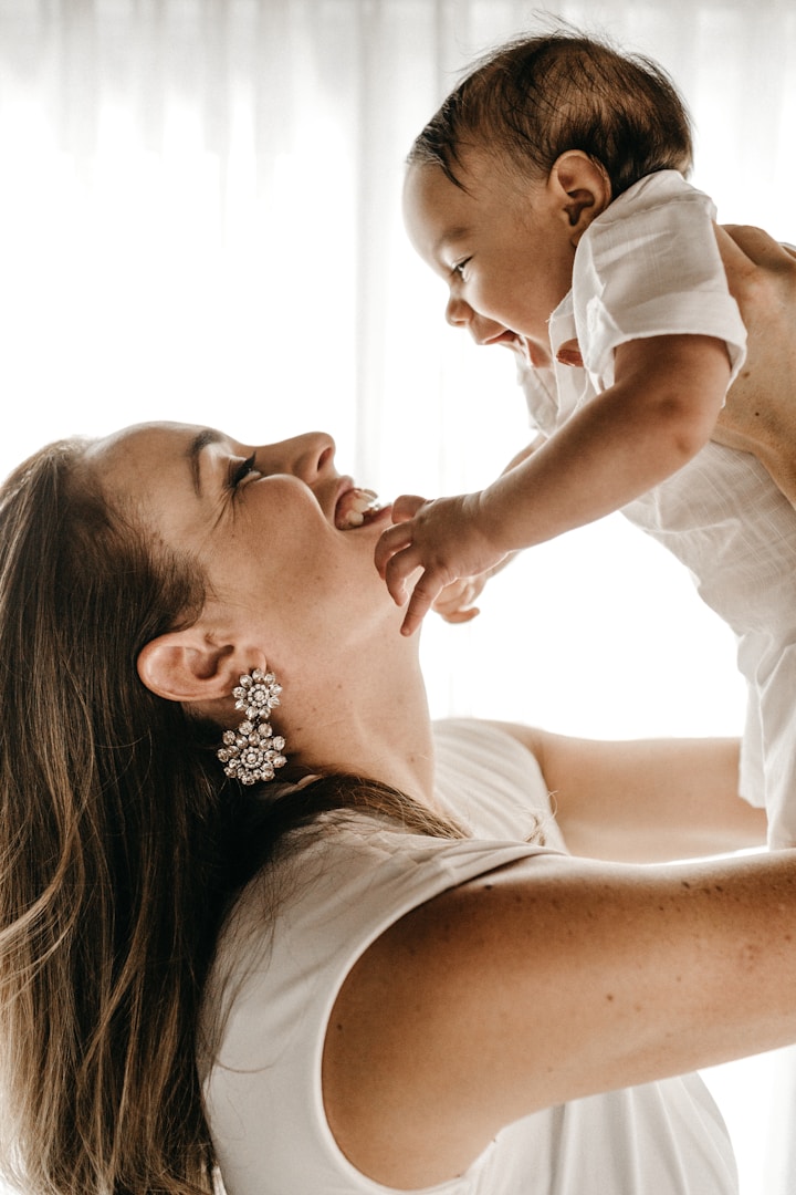 A mother holds her baby, both smiling at each other in a bright, soft-lit setting.