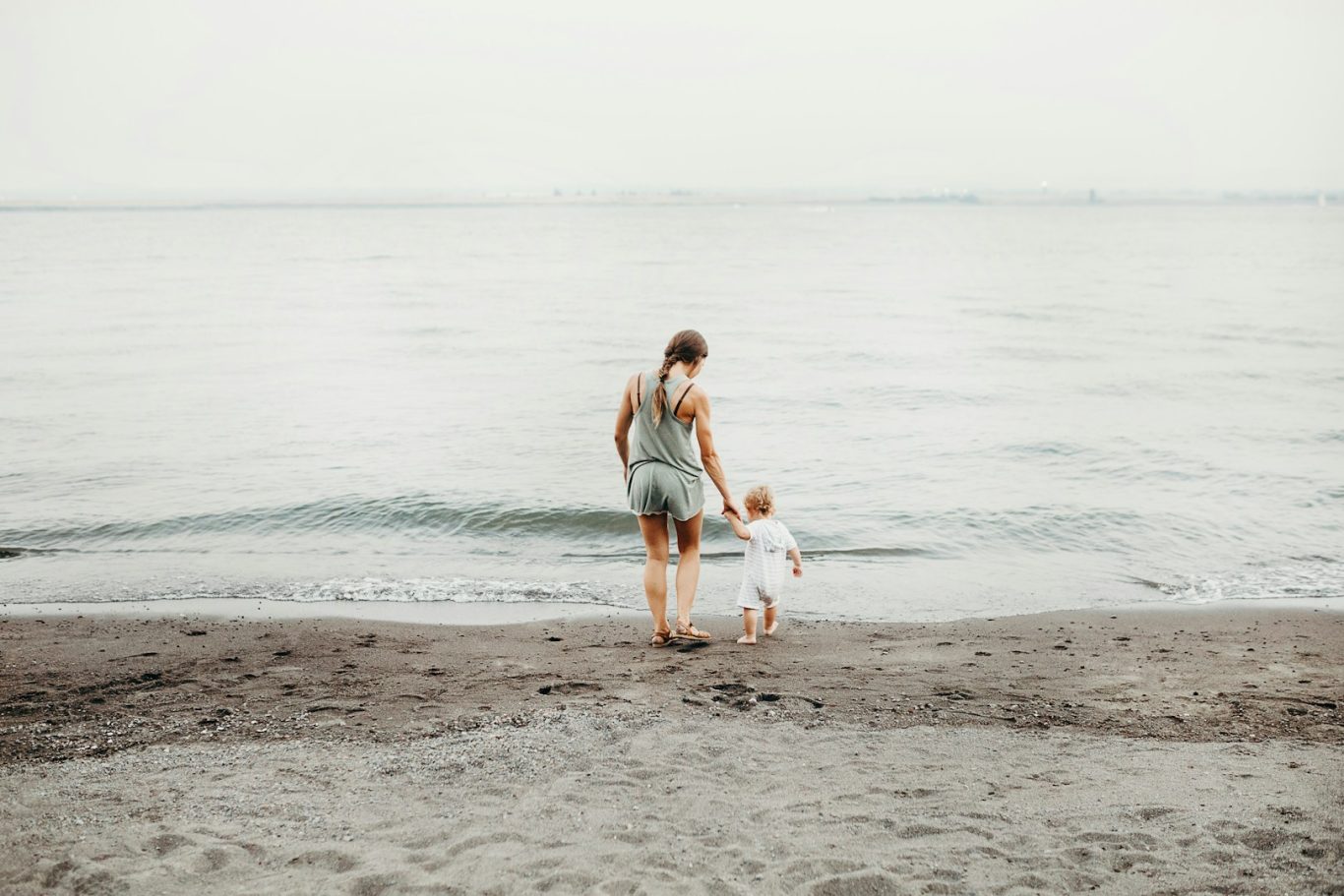 A person and child walking hand-in-hand along a sandy beach by the water's edge.